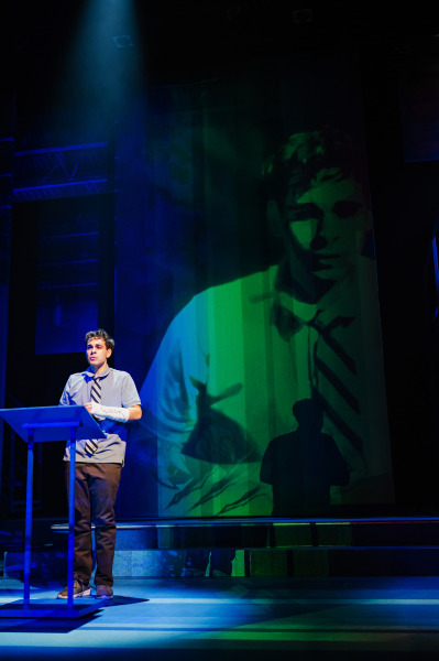 In Dear Evan Hansen, a teenage boy stands at a podium under a narrow spotlight, delivering a speech. Behind him, a large semi-transparent projection of his face fills the stage, casting layered shadows and emphasizing his vulnerability and inner thoughts.