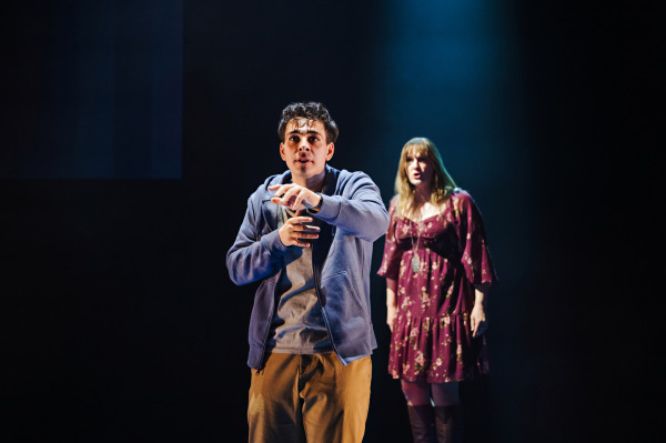 A young man stands downstage in Dear Evan Hansen, reaching forward with urgency, his expression intense. Behind him, a young woman in a patterned dress stands slightly out of focus, reacting with concern as dramatic lighting isolates the pair against a dark background.