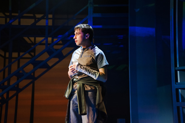A teenage boy with a cast labelled “Connor” stands in profile under moody blue and amber lighting in Dear Evan Hansen. His posture is tense and inward, framed by industrial-style set pieces and stairs behind him.