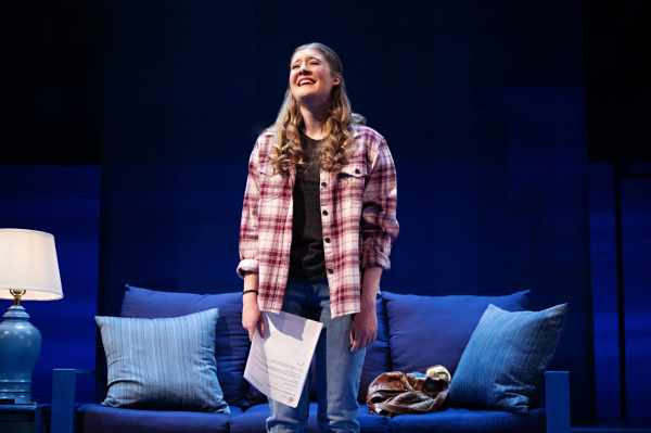 A young woman stands centre stage in Dear Evan Hansen, singing with emotion while holding papers in her hand. She is positioned in front of a couch and table lamps that suggest a living room setting, softly lit in cool tones.