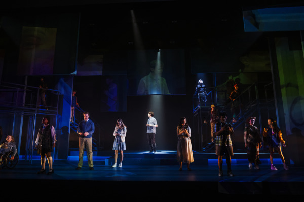  A young man stands alone in a tight beam of light at centre stage in Dear Evan Hansen, while other characters occupy elevated platforms around him. Large projected images hover above, reinforcing a feeling of isolation within a connected digital world.