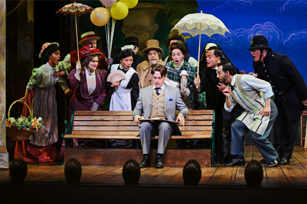 In a lively park scene from Finding Neverland, a well-dressed man sits on a wooden bench reading as a group of Victorian-era characters gathers around him in animated curiosity. The ensemble wears period costumes including hats, parasols, and layered dresses, leaning in with surprised and delighted expressions. Brightly painted scenery featuring trees and classical statues creates a whimsical, storybook setting.