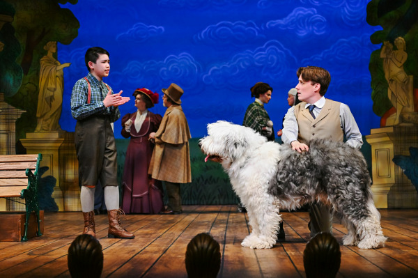 A young performer stands facing an adult in Finding Neverland, gesturing earnestly while speaking. Beside them stands a large, fluffy dog puppet operated at waist height, its textured fur and expressive stance adding a playful theatrical element. Behind them, other characters in period costumes move across the stage, set against a painted sky backdrop with stylized clouds, creating a light and imaginative atmosphere.