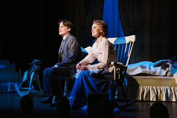 Two performers sit side by side onstage in Finding Neverland, lit by a warm spotlight against a darkened backdrop. One wears a tailored suit, while the other is dressed in a soft, period-style blouse and long skirt, seated in a wooden rocking chair. Their expressions are gentle and reflective as they look outward, suggesting a quiet, emotional moment. Behind them, a child lies resting in a bed, partially illuminated, adding a sense of intimacy and tenderness to the scene.