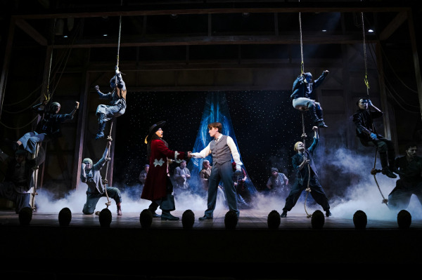 A dramatic, high-energy scene from Finding Neverland features performers climbing and swinging on ropes across a large wooden stage structure. At centre, two characters face one another, clasping hands in a moment of connection, while surrounding ensemble members strike bold, action-filled poses. Smoke effects and star-like lighting in the background create a sense of adventure and fantasy, evoking the imaginative world of Neverland.