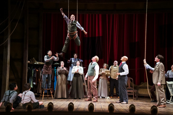 A dynamic stage moment from Finding Neverland shows a performer suspended mid-air by ropes, lifted above a group of onlookers who gaze upward in amazement. The cast, dressed in period costumes, stands beneath red theatre curtains as the airborne figure appears to fly. The composition captures a sense of wonder and theatrical magic as the ensemble reacts to the spectacle.