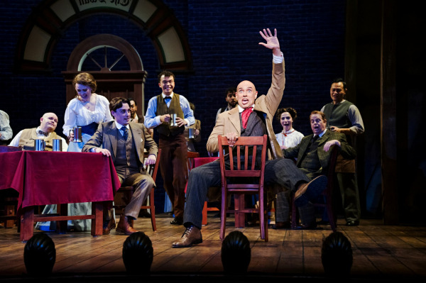 A comedic ensemble moment unfolds in Finding Neverland as a bald performer sits dramatically on a chair at centre stage, raising one arm high with exaggerated flair. Around him, cast members in period attire react with laughter and surprise, holding mugs and leaning into the moment. The set resembles a warmly lit interior space with brick walls and wooden furniture, enhancing the lively, theatrical energy.