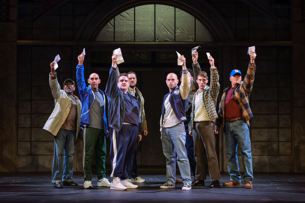 Seven men stand shoulder to shoulder on stage in The Full Monty at Huron Country Playhouse, each raising an envelope high above their heads in a powerful moment of solidarity. Dressed in everyday clothing ranging from work jackets and track suits to jeans and flannel shirts, the performers face the audience with determined expressions beneath warm theatrical lighting, framed by the entrance of an industrial-style building that reflects the show's working-class setting.