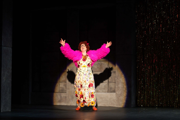 A performer stands centre stage in a spotlight during The Full Monty at Huron Country Playhouse, wearing a bright pink feathered jacket over a long floral dress with bold jewellery and red shoes. With arms outstretched and a joyful expression, the performer commands the stage beside a shimmering tinsel curtain, while the darkened industrial-style set draws focus to the colourful costume and theatrical pose.
