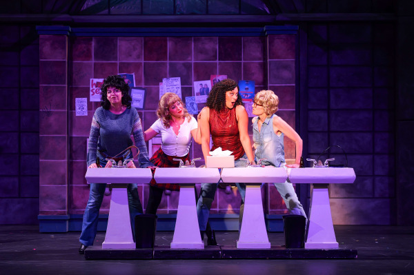 Four women gather around a row of washroom sinks during a lively scene from The Full Monty at Huron Country Playhouse. Leaning in with animated expressions as if sharing gossip, the performers wear casual everyday clothing ranging from jeans and sleeveless tops to a sparkling red blouse. Set against a tiled washroom backdrop covered with posters and notices, the scene is lit with vibrant purple and pink stage lighting that highlights the show's humour and camaraderie.