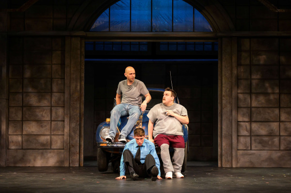 Three men gather around the hood of a battered blue car during a scene from The Full Monty at Huron Country Playhouse. One performer in a security guard uniform sits on the stage floor while two others perch on the car, dressed in casual T-shirts, jeans, and sweatpants. Their thoughtful expressions suggest an important conversation as warm stage lighting illuminates the industrial building façade behind them, emphasizing the show's working-class setting and themes of friendship and resilience.
