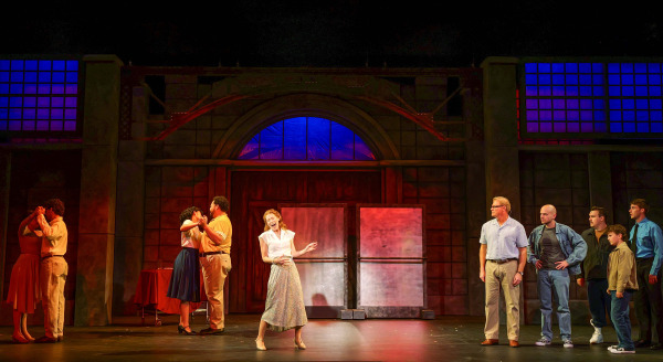 A woman sings at centre stage during a scene from The Full Monty at Huron Country Playhouse. On stage left, two couples slow dance together, while on stage right a group of five men, a young boy, and a security guard watch from across the stage. Warm amber and cool blue lighting illuminate the industrial-style set, with large doors and factory windows framing the ensemble.