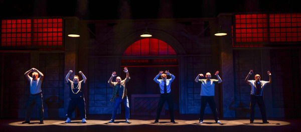 Six performers stand evenly spaced across the stage during The Full Monty at Huron Country Playhouse, each striking a playful dance pose with their arms raised. Individual spotlights illuminate the performers against an industrial-style set, while dramatic red and blue lighting fills the large windows behind them.