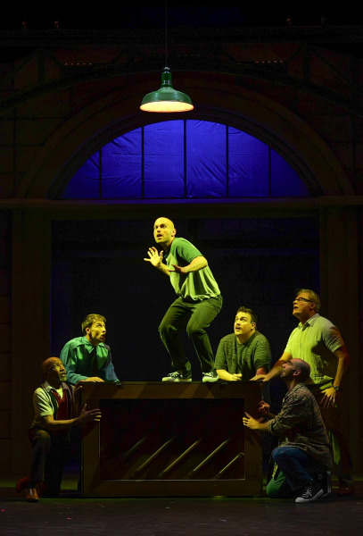 Six men gather around an upright piano during a dramatic scene from The Full Monty at Huron Country Playhouse. One performer crouches on top of the piano beneath a single hanging industrial light while the others kneel or stand around him, looking up with anxious expressions. The industrial-style set is bathed in moody blue and green lighting, drawing focus to the tense moment at centre stage.