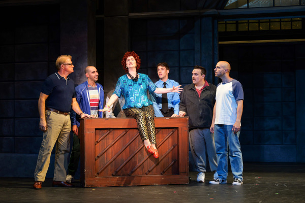 A woman with curly red hair sits atop an upright piano, singing with her arms outstretched during a scene from The Full Monty at Huron Country Playhouse. Gathered around the piano, five men and a security guard watch her performance with attentive expressions. The performers wear casual everyday clothing, and the industrial-style set is illuminated with warm stage lighting against dark steel walls.