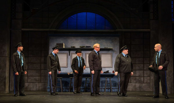 Six performers dressed as security guards stand across the stage during a scene from The Full Monty at Huron Country Playhouse. Wearing matching black uniforms with blue shirts and peaked caps, the group exchanges amused and animated expressions in front of a security office set with lockers, stools, and a counter. Warm stage lighting highlights the performers against the industrial-style backdrop.