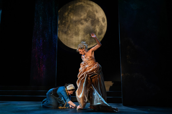 In Into the Woods at St. Jacobs Country Playhouse, a performer in an elaborate shimmering gold gown stands beneath a huge projected full moon, one arm raised dramatically while the other gathers the voluminous skirt. At their feet, another performer in a blue dress and headscarf kneels on the stage, reaching toward a gold shoe. The scene is framed by deep black scenery and cool theatrical lighting.