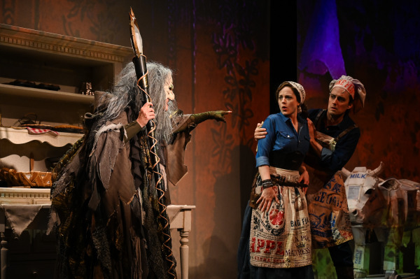 In Into the Woods at St. Jacobs Country Playhouse, the Witch stands at left in a tattered brown costume and long grey hair, pointing accusingly across the stage while holding a tall staff. At right, two performers dressed as the Baker and Baker’s Wife cling together with startled expressions. A whimsical cow prop made from printed paper and packaging stands beside them, while warm orange and purple lighting and shadowy forest projections fill the background.