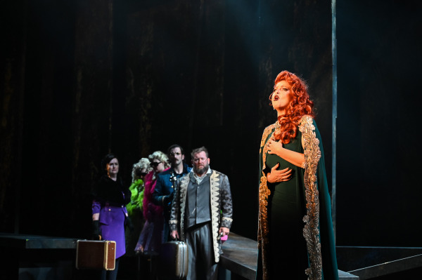 In Into the Woods at St. Jacobs Country Playhouse, a red-haired performer in a dark green gown with ornate gold trim stands in a strong spotlight at the front of the stage, singing with one hand pressed to their chest and the other across their stomach. Several performers stand in shadow behind them wearing a mix of formal and colourful costumes, some holding suitcases, as the dark forest set fades into the background.