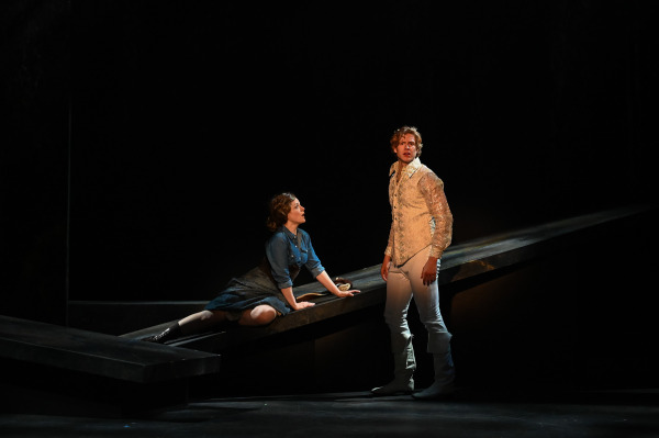 In Into the Woods at St. Jacobs Country Playhouse, two performers share a quiet moment on the darkened forest set. One sits on a sloped platform wearing a blue dress and looks upward toward another performer standing nearby in an ornate cream-coloured jacket, light trousers, and boots. A focused pool of light isolates the pair against an otherwise black stage.