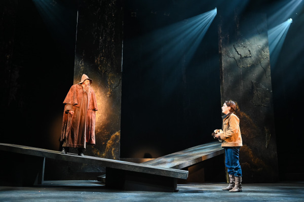 In Into the Woods at St. Jacobs Country Playhouse, a hooded performer in a long brown cloak stands elevated on a slanted platform at left, facing a younger performer across the stage. The younger performer wears a tan jacket, blue trousers, and brown boots and holds an object close to their chest. Tall, distressed scenic panels rise behind them as cool beams of light cut through the dark, atmospheric set.