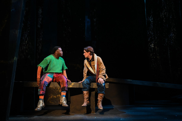 In Into the Woods at St. Jacobs Country Playhouse, two performers sit side by side on the edge of a low platform in the shadowy forest setting. One wears a bright green crocheted top, colourful patchwork trousers, striped socks, and silver boots, while the other wears a tan jacket, scarf, blue trousers, and brown boots. They lean toward one another in conversation beneath a narrow pool of warm stage light.