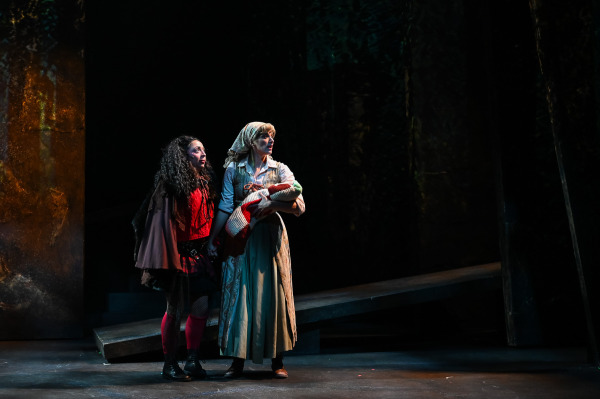 In Into the Woods at St. Jacobs Country Playhouse, two performers stand close together in the dark forest setting, both looking upward with apprehensive expressions. One wears layered black and red clothing with long dark curls, while the other wears a pale blue and cream dress with a headscarf and cradles a bundled infant in a red, white, and green knitted blanket. A sloped platform and towering dark scenic panels recede behind them.