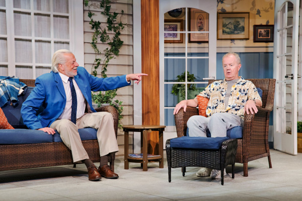 In Jonas & Barry in the Home, a performer seated on a wicker sofa leans forward with a broad smile and points across the stage toward another performer. The second performer sits stiffly in a wicker armchair, looking surprised, dressed in a patterned shirt and grey pants. A small wooden table and ottoman sit between them, surrounded by the windows, French doors, greenery, and warm-toned furnishings of the patio set.
