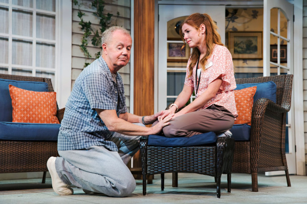 In Jonas & Barry in the Home, a performer in a grey plaid shirt and grey pants kneels beside a wicker ottoman, gently holding the hands of a red-haired performer seated cross-legged on the cushion. The seated performer, dressed in a pink floral blouse, brown pants, white sneakers, and an identification lanyard, looks emotional as the two share a quiet moment. Wicker seating with blue cushions, orange pillows, white-framed windows, and an open doorway surround them.