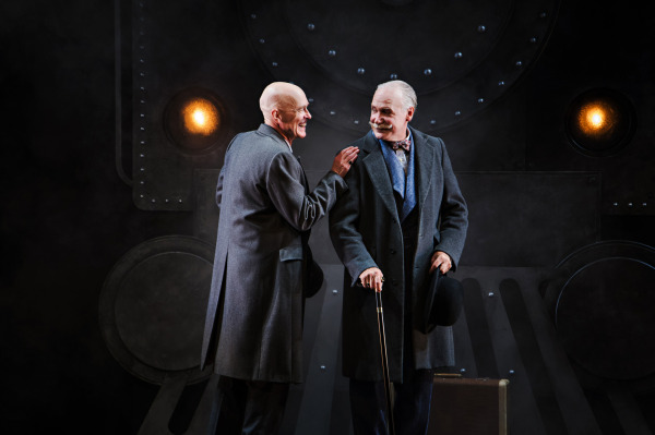 Two performers stand facing one another in front of a large, industrial-style train set with circular metal panels, riveted surfaces, and glowing amber lights. Both wear dark formal suits, with one holding a walking cane and resting a foot on a brown suitcase. The performers smile and gesture toward each other against the smoky, grey-toned backdrop, illuminated by warm stage lighting.