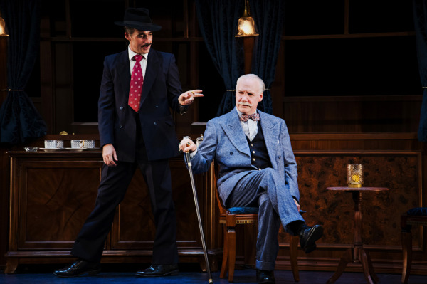 Inside an elegant, wood-panelled train carriage, a performer wearing a dark three-piece suit, red patterned tie, and black fedora stands gesturing toward another performer seated nearby. The seated performer wears a light blue suit, holds a walking cane, and sits with one leg crossed while raising a hand. Ornate wooden cabinetry, a small round table, upholstered chairs, dark blue curtains, and hanging brass lamps complete the luxurious period setting, illuminated by warm amber lighting.