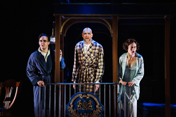 Three performers stand together behind a decorative black-and-gold railing on a raised section of the train set. The performer on the left wears a dark striped robe and grips the doorway, while the central performer wears a yellow-and-brown plaid dressing gown and faces forward with an intense expression. On the right, a performer in a pale blue-green robe raises a hand toward their mouth in apparent surprise. A rounded archway frames the group, while dramatic blue and amber lighting creates a shadowy atmosphere.