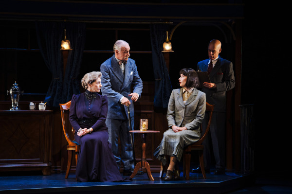 Four performers gather inside a richly decorated train carriage. A performer in a long purple dress sits at the left, while another in a pale green-grey suit sits at the right, looking toward a white-haired performer leaning forward between them. A fourth performer stands behind, dressed in a dark suit and holding a small notebook. Polished wooden cabinetry, upholstered chairs, brass hanging lamps, and dark curtains establish the elegant period interior, with warm lighting illuminating the group against the darker background.