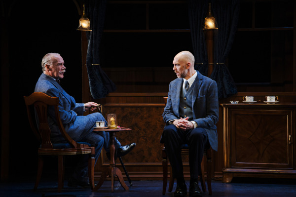 Two performers sit facing one another inside an elegant train carriage, separated by a small wooden table holding a silver tea service. The performer on the left wears a blue suit and sits with legs crossed, leaning back in an upholstered chair, while the performer opposite wears a grey three-piece suit and leans forward with hands clasped. Ornate wood-panelled cabinets, dark curtains, brass pendant lights, and neatly arranged white teacups frame the conversation, with warm lighting focused on the performers against a dimly lit backdrop.
