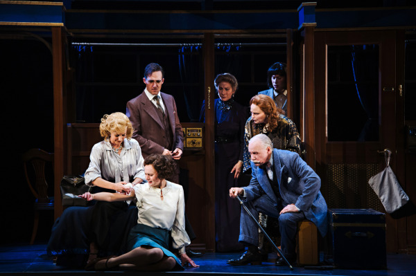 A group of performers gathers around a person seated on the floor of an elaborate train carriage. One performer kneels beside the seated figure, appearing to offer assistance, while another crouches nearby holding a walking cane. Additional performers stand behind them, looking toward the centre of the scene. The costumes include formal suits, tailored dresses, and period accessories in muted blue, grey, cream, and brown tones. Ornate wooden compartment walls, dark blue curtains, brass lamps, and scattered luggage establish the train setting, while dramatic lighting concentrates attention on the group.