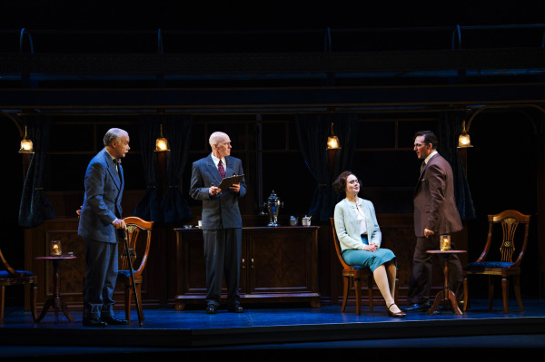 Four performers are positioned across the stage inside a dimly lit train carriage. At centre, a performer in a dark suit stands holding a small notebook or papers, while another in a blue suit stands to the left. A seated performer wearing a light blue-green outfit looks toward a figure on the right, dressed in a brown suit and gesturing during the conversation. Wooden chairs, a long polished cabinet, a silver tea service, dark curtains, and hanging brass lamps create the period train interior, illuminated by pools of warm light.