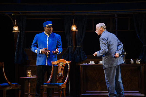 Two performers face one another inside an elegant train carriage. On the left, a performer dressed as a train attendant wears a vivid royal blue uniform with gold trim, brass buttons, and a matching cap, gesturing with both hands. Opposite, a performer in a grey-blue suit leans forward with hands clasped behind their back. Ornate wooden chairs, a polished cabinet with white teacups and silver serving pieces, dark curtains, and brass pendant lamps fill the background, with warm lighting highlighting the exchange.