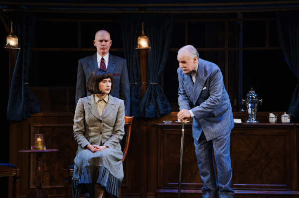 Three performers gather inside an elegant, wood-panelled train carriage. At centre, a performer wearing a tailored grey-green skirt suit sits upright in a wooden chair with hands folded in their lap. Behind them stands a performer in a dark suit and red tie, while another in a light blue suit leans forward on a walking cane, appearing to address the seated figure. Brass pendant lamps, dark blue curtains, wooden cabinets, and a silver tea service frame the scene, with warm stage lighting illuminating the performers.