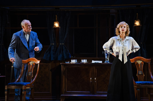 Two performers stand inside a richly detailed train carriage. On the left, a white-haired performer in a blue suit rests one hand on the back of an ornate wooden chair and gestures with the other. On the right, a performer with shoulder-length wavy blonde hair stands with hands on hips, wearing a pale blue blouse with a bow at the neck and a long black skirt. A polished wooden cabinet topped with white teacups and saucers, dark curtains, and glowing brass pendant lamps establish the elegant period setting, while focused lighting emphasizes the performers against the darkened stage.