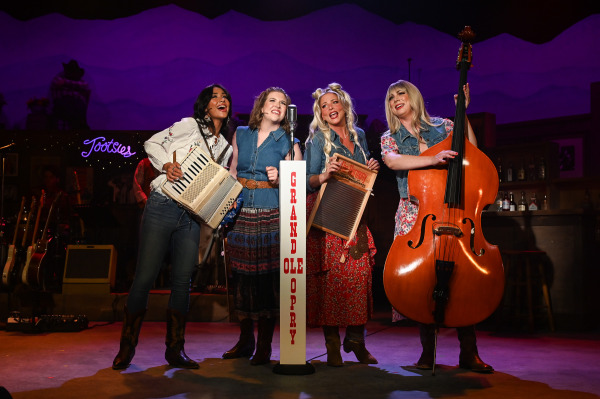 Four performers gather around a vintage-style microphone stand labelled “Grand Ole Opry” during Nashville Music City. One performer plays an accordion, another stands at centre singing, a third holds a washboard, and a fourth plays an upright bass. Dressed in country-inspired outfits featuring denim, floral prints, and western boots, the group harmonizes under warm stage lighting. The rustic honky-tonk set includes guitars, bar shelving, and a glowing “Tootsies” sign in the background, evoking the atmosphere of a classic Nashville country music performance.