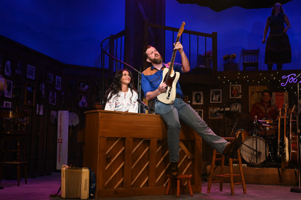 A guitarist balances on the edge of an upright piano while playing an electric guitar during an energetic musical number in Nashville Music City. A pianist smiles up from the keyboard below as other musicians perform nearby. The country music venue set features wood finishes, vintage photographs, and warm stage lighting that enhances the playful concert atmosphere.