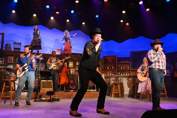 Two country music performers sing at the front of the stage during a high-energy ensemble number in Nashville Music City. A guitarist, bassist, and additional musicians accompany them while performers dance on an elevated platform above. Colourful concert lighting and western-inspired costumes create the feel of a lively Nashville stage show.