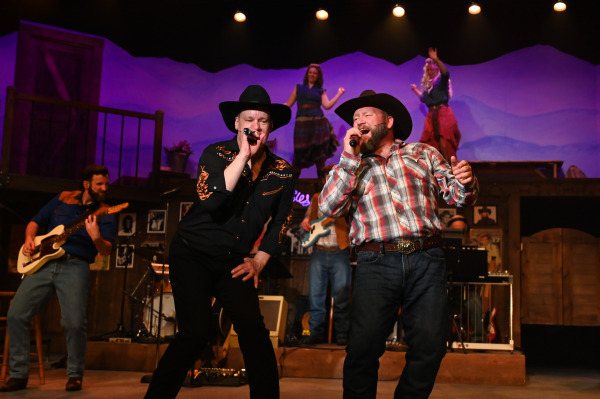 A performer passionately sings into a handheld microphone at centre stage in Nashville Music City, leaning back as if hitting a powerful note. Nearby, an accordion player and guitarist accompany the song while additional performers watch from an elevated balcony. Warm amber lighting and rustic country décor reinforce the show’s celebration of Nashville’s musical heritage.
