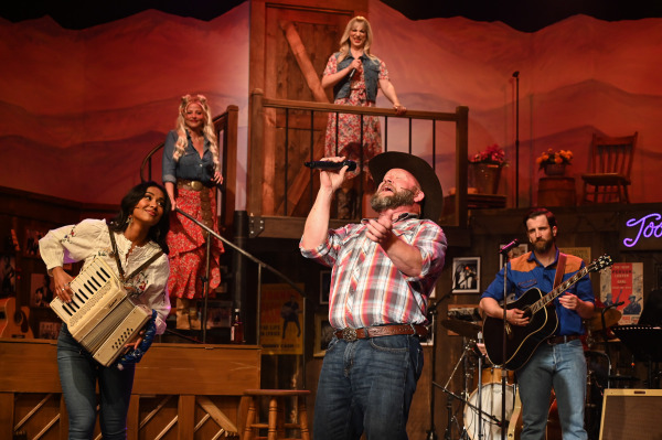 A performer in a plaid western shirt and black cowboy hat sings passionately into a handheld microphone at centre stage during Nashville Music City. An accordion player stands to one side, smiling toward the singer, while a guitarist accompanies the performance nearby. Two additional performers in floral dresses and denim vests watch from an elevated balcony above, framed by rustic wooden railings and a barn-style door. Warm amber lighting bathes the country music venue set, which features a piano, vintage posters, and honky-tonk décor, creating the atmosphere of a lively Nashville concert.