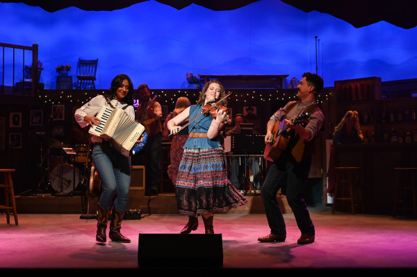 Three performers share the spotlight during an energetic musical number in Nashville Music City. One performer plays accordion, another performs a fiddle solo, and a third strums an acoustic guitar as they smile and interact with one another. Musicians and singers fill the background, while blue and purple stage lighting washes over the country-themed set, creating the feel of an intimate live music venue.