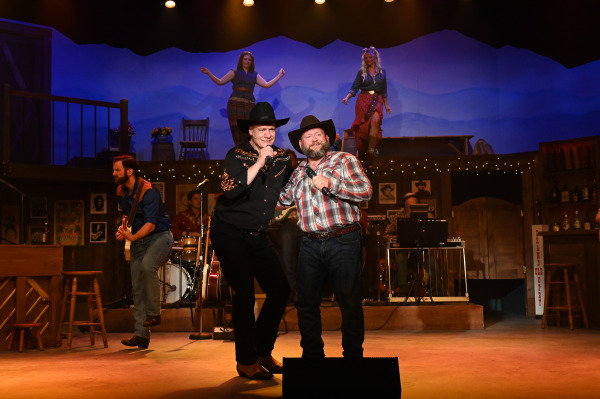 Two performers share a duet at centre stage in Nashville Music City, singing into handheld microphones while standing close together. Behind them, fellow performers dance on an elevated platform and musicians play guitar, bass, and drums. The rustic honky-tonk set, accented with warm lighting and country music memorabilia, evokes the spirit of a Nashville music hall.