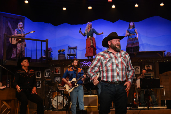 A country music performance unfolds on the stage of Nashville Music City as a bearded performer in a cowboy hat sings into a handheld microphone at centre stage. Around him, musicians play guitar, drums, and keyboards while three performers sing and dance from an elevated balcony. The set resembles a rustic country music venue, complete with framed photographs, wood panelling, and a glowing “Tootsies” sign. Warm amber and blue lighting create the atmosphere of a lively Nashville concert.