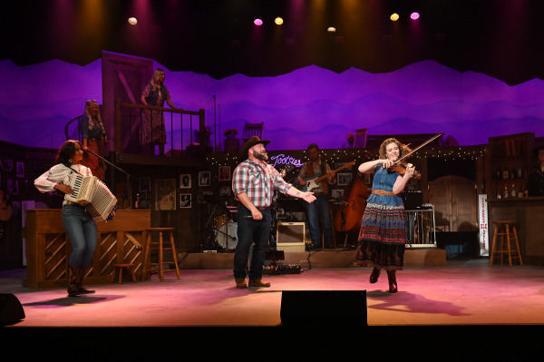 A fiddle player takes centre stage during a spirited instrumental feature in Nashville Music City. An accordion player and vocalist look on as the musician performs, while the live band accompanies from the background. Purple and amber lighting illuminate the stage, highlighting the energy and skill of the featured performance.