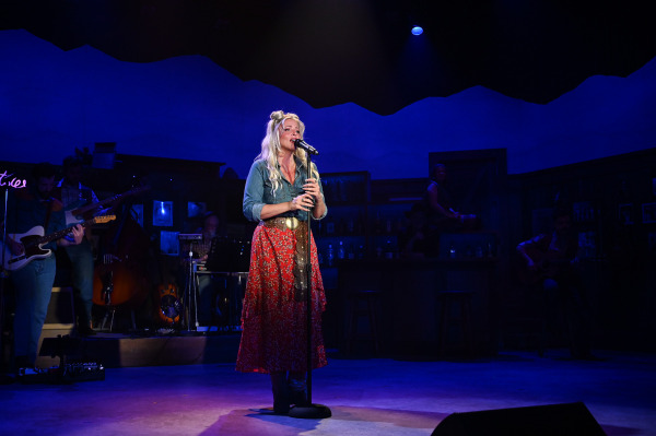 A performer stands alone at a microphone, singing a heartfelt ballad in Nashville Music City. Dressed in a flowing red floral skirt and denim shirt, they are illuminated by a soft spotlight against a backdrop of deep blue lighting. Members of the live band are partially visible in the shadows, emphasizing the intimate and emotional focus of the solo performance.