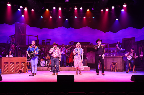 The full company of Nashville Music City performs together during a vibrant ensemble number. Singers stand across the front of the stage with handheld microphones while musicians play behind them on guitar, bass, drums, banjo, and piano. Bright pink, purple, and amber stage lighting fills the theatre, highlighting the energetic atmosphere of a country music concert.